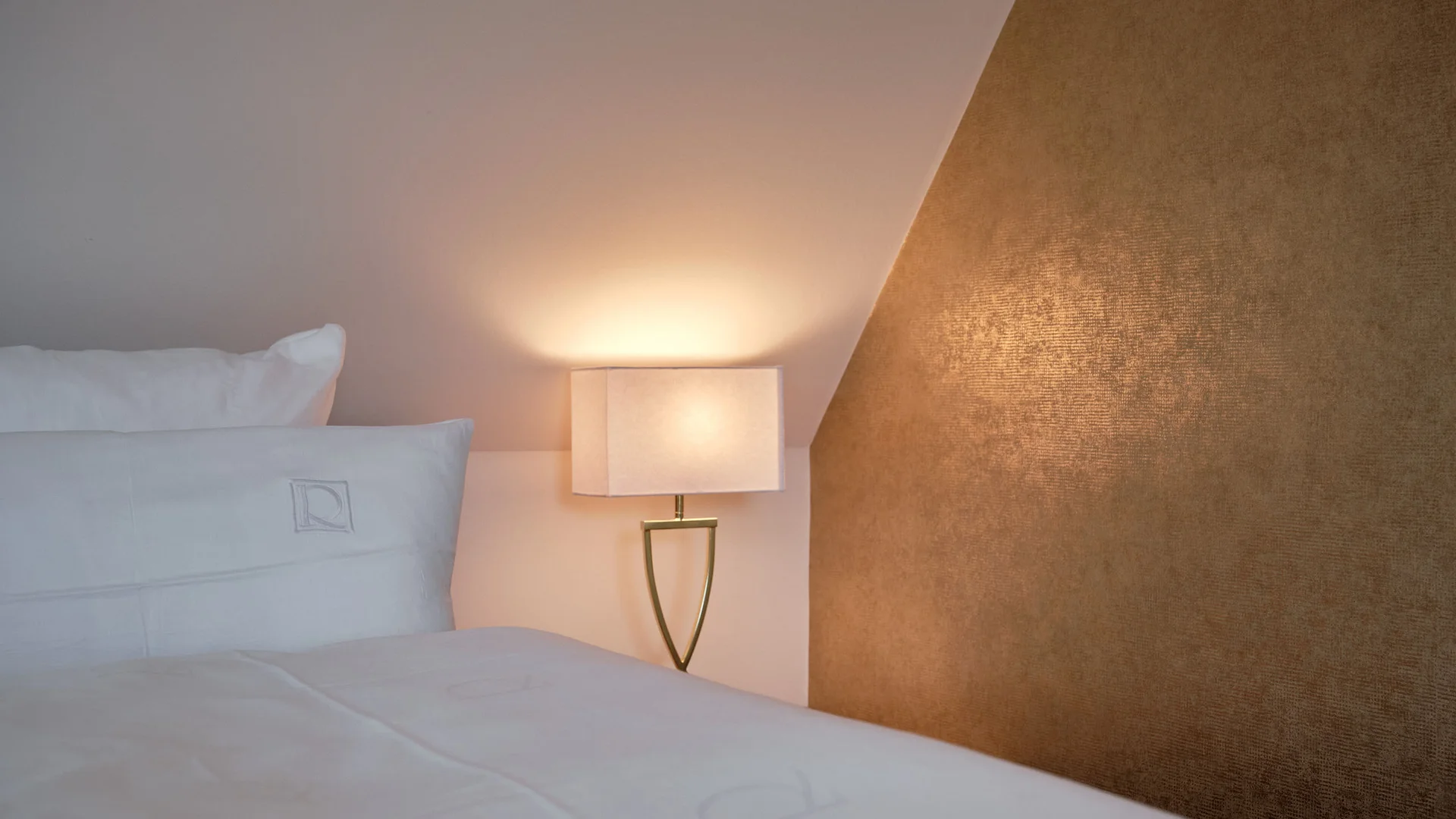 Weißes Bett mit Kissen und eine beleuchtete Wandlampe an goldener Wand