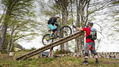 Mountainbiker fährt über hölzerne Rampe, begleitet von einem Helfer im Wald