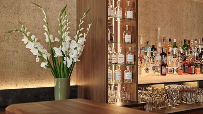 Bar mit Holztheke, weißen Blumen und beleuchteten Regal mit Spirituosen und Gläsern