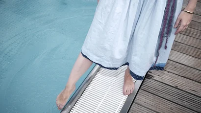 Frau im Kleid steht barfuß am Poolrand auf Holzdeck