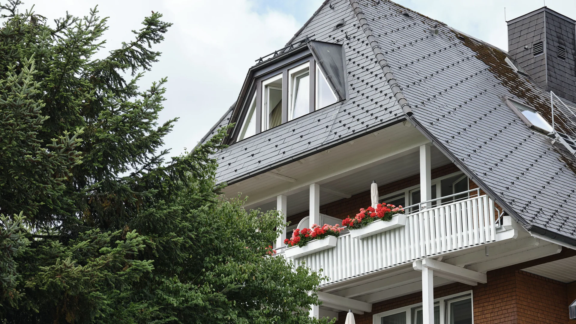 Haus mit grauem Schieferdach und weißen Balkonen mit roten Blumen
