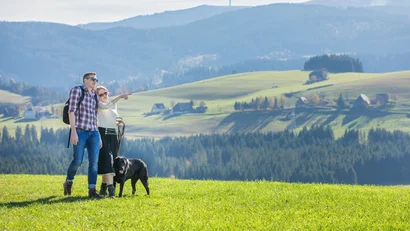 Paar mit Hund wandert durch grüne Wiesen mit Bergen im Hintergrund