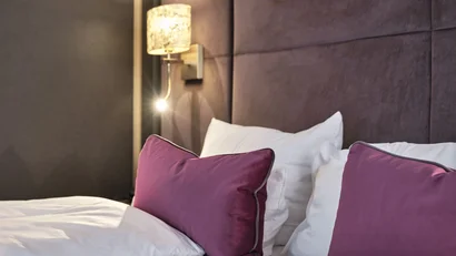 Modernes Bett mit weißen Laken und violetten Kissen vor gepolstertem Kopfteil